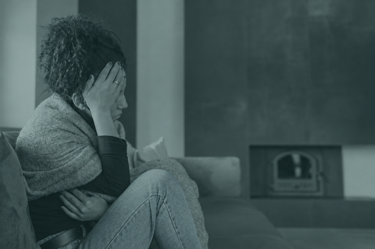 A tired woman with curly hair, wrapped in a grey knit shawl, sits on a sofa with one hand on her forehead and the other arm across her body