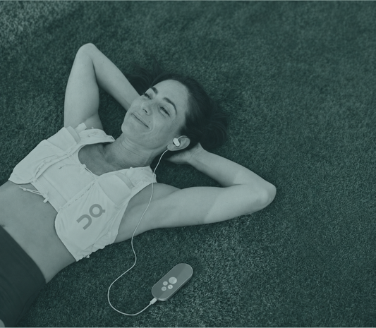 Woman lying on grass with eyes closed, wearing the Nurosym ear clip with the device resting on her torso