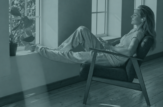 Woman relaxing in armchair by window wearing a Nurosym ear piece while using her phone
